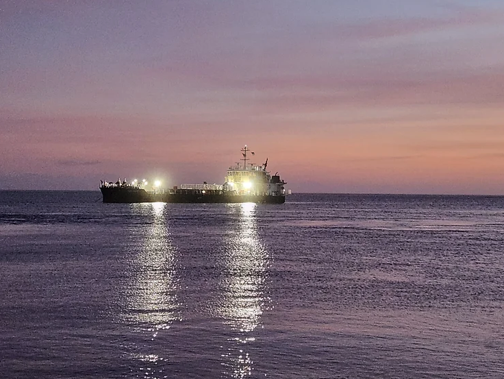 An image of a large dredging ship illuminated with lights sits in the distance on the Pamlico Sound. the water and sky are colored lavender, with hints of orange and pink above the vessel.